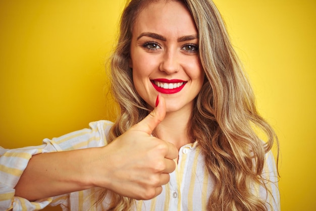 Free photo young beautiful woman wearing stripes shirt standing over yellow isolated background happy with big smile doing ok sign thumb up with fingers excellent sign
