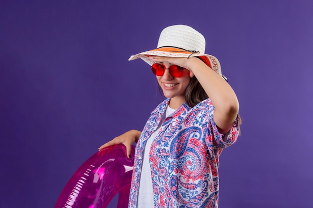 Young beautiful woman wearing summer hat and red sunglasses holding inflatable ring looking far away with hand to look something smiling and happy, ready to holiday concept over purple