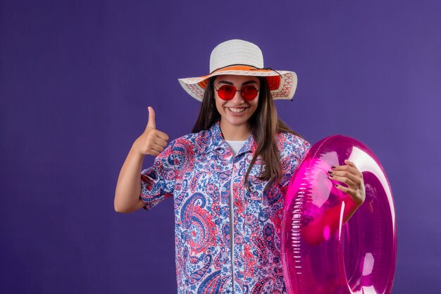 Young beautiful woman wearing summer hat and red sunglasses holding inflatable ring wit happy face smiling cheerfully showing thumbs up over purple wall