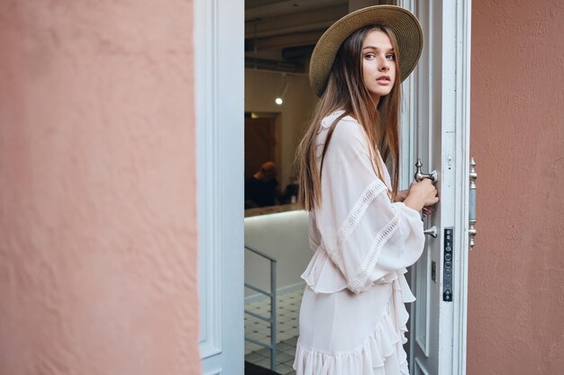 Young beautiful woman in white dress and hat thoughtfully looking in camera while opening white door of cafe