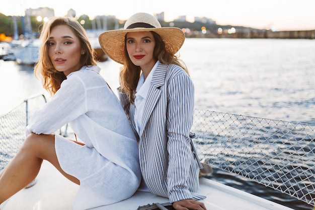 白いシャツを着た若い美しい女性とストリップジャケットと麦わら帽子のゴージャスな女性が背景に素晴らしい港の景色を望むヨットで一緒にカメラで夢のように見ています