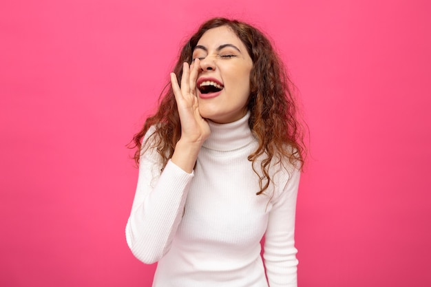 Young beautiful woman in white turtleneck shouting or calling holding hand over mouth standing over pink wall