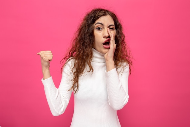 Young beautiful woman in white turtleneck telling a secret holding hand on mouth pointing with thumb to the side standing on pink