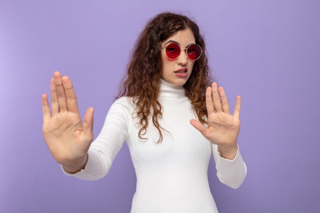 Young beautiful woman in white turtleneck wearing red glasses worried making stop gesture with hands standing on purple