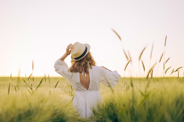 밀짚모자를 쓰고 흰 드레스를 입은 금발의 긴 머리를 한 젊은 아름다운 여성이 밀밭에서 꽃을 모은다. 태양, 여름에 비행 머리. 몽상가를 위한 시간, 황금빛 일몰.