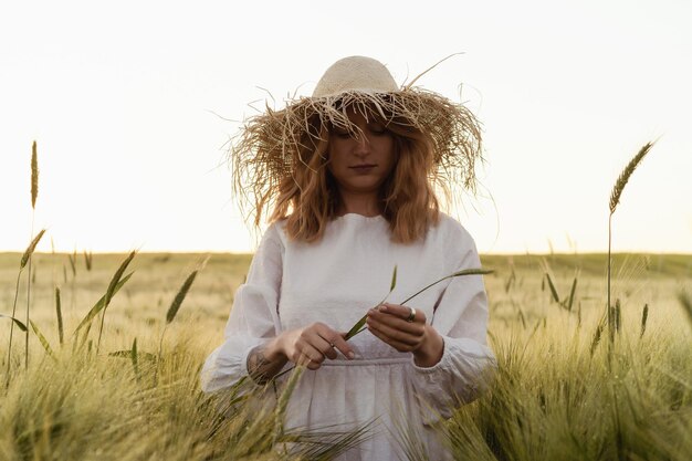 麦わら帽子の白いドレスを着た金髪の長い髪の若い美しい女性は、麦畑に花を集めます。太陽の下で飛んでいる髪、夏。夢想家のための時間、黄金の夕日。