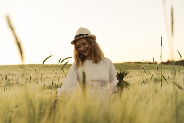 밀짚모자를 쓰고 흰 드레스를 입은 금발의 긴 머리를 한 젊은 아름다운 여성이 밀밭에서 꽃을 모은다. 태양, 여름에 비행 머리. 몽상가를 위한 시간, 황금빛 일몰.