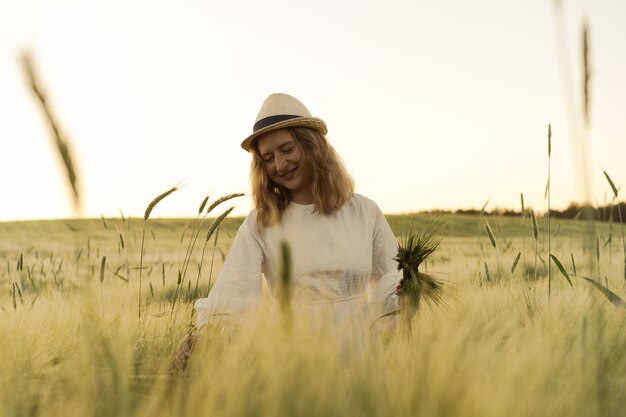 молодая красивая женщина с длинными светлыми волосами в белом платье в соломенной шляпе собирает цветы на пшеничном поле. Летающие волосы на солнце, лето. Время мечтателей, золотой закат.