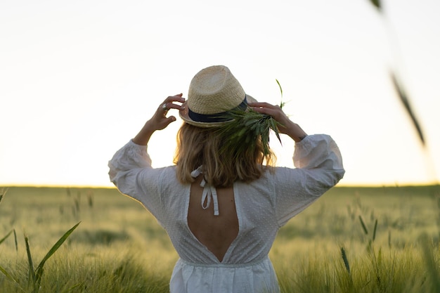 麦わら帽子の白いドレスを着た金髪の長い髪の若い美しい女性は、麦畑に花を集めます。太陽の下で飛んでいる髪、夏。夢想家のための時間、黄金の夕日。