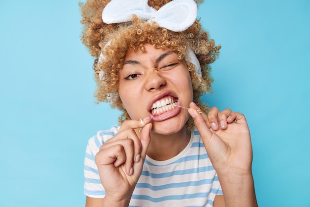 巻き毛の櫛の髪を持つ若い美しい女性は、歯をきれいにするためにデンタルフロスを使用してヘッドバンドと青い背景の上に分離されたカジュアルなストライプのTシャツを着用します歯のフロス歯のケアルーチンのコンセプト