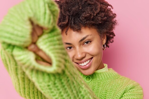 Young beautiful woman with curly hair has playful expression smiles pleasantly at camera wears long sleeved knitted sweater isolated over pink background. Sincere emotions and feelings concept