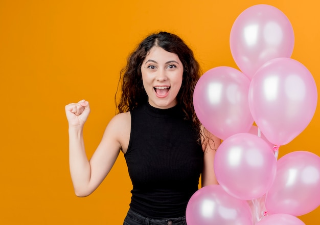 Молодая красивая женщина с вьющимися волосами держит кучу воздушных шаров, сжимая кулак, счастливая и взволнованная концепция вечеринки по случаю дня рождения, стоящая над оранжевой стеной