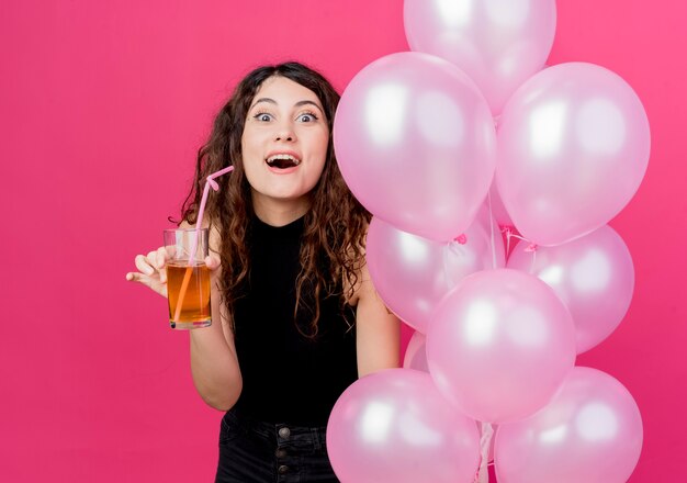 Молодая красивая женщина с вьющимися волосами, держащая кучу воздушных шаров и коктейль, выглядит удивленной, весело улыбаясь, стоя над розовой стеной