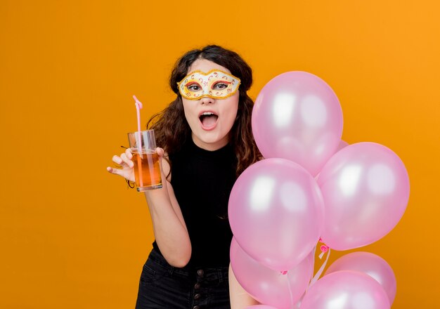 Young beautiful woman with curly hair holding bunch of air balloons and cocktail in party mask happy and cheerful birthday party concept over orange