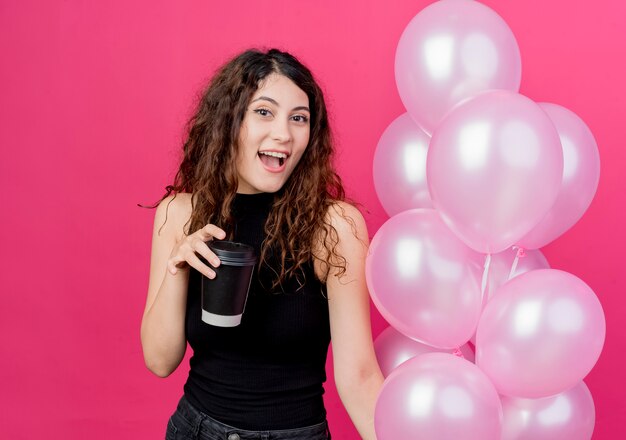 Young beautiful woman with curly hair holding bunch of air balloons and coffee cup smiling cheerfully standing over pink wall