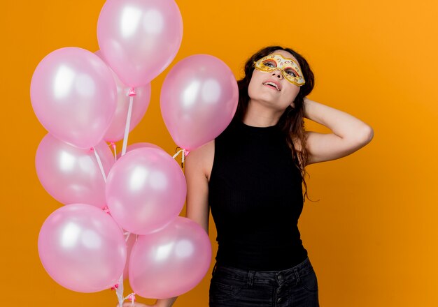 Young beautiful woman with curly hair holding bunch of air balloons looking up happy and cheerful birthday party concept standing over orange wall