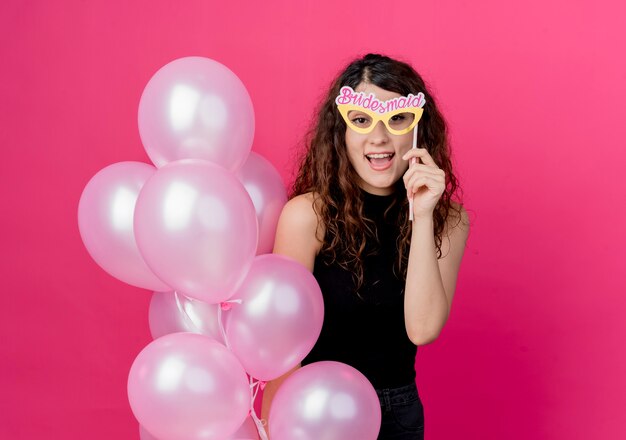 Young beautiful woman with curly hair holding bunch of air balloons and paper glasses smiling cheerfully standing over pink wall