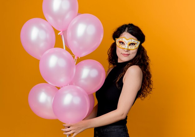 Young beautiful woman with curly hair holding bunch of air balloons in party mask happy and cheerful birthday party concept  over orange