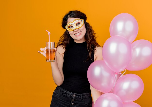 Young beautiful woman with curly hair holding bunch of air balloons in party mask happy and cheerful drinking cocktail birthday party concept standing over orange wall