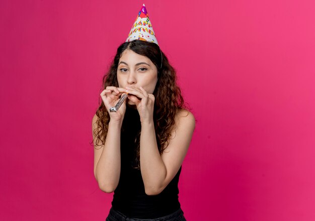 Молодая красивая женщина с вьющимися волосами в праздничной кепке, дует в свисток, счастливая и позитивная концепция вечеринки по случаю дня рождения, стоящая над розовой стеной