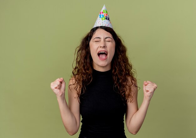 Young beautiful woman with curly hair in a holiday cap clenching fists happy and exited standing over light wall