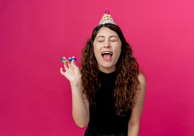 Young beautiful woman with curly hair in a holiday cap crazy happy birthday party concept standing over pink wall