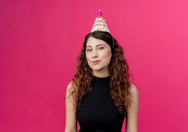 ピンクの壁の上に立っている休日の帽子幸せでポジティブな笑顔の誕生日パーティーのコンセプトで巻き毛の若い美しい女性
