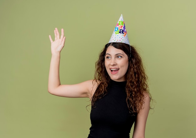 Молодая красивая женщина с вьющимися волосами в праздничной шапочке счастлива и позитивно машет рукой, стоя над светлой стеной