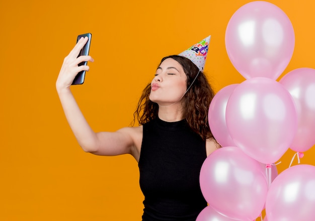 Молодая красивая женщина с вьющимися волосами в праздничной шапочке держит воздушные шары, дует поцелуй, принимая селфи-концепцию вечеринки по случаю дня рождения, стоя над оранжевой стеной