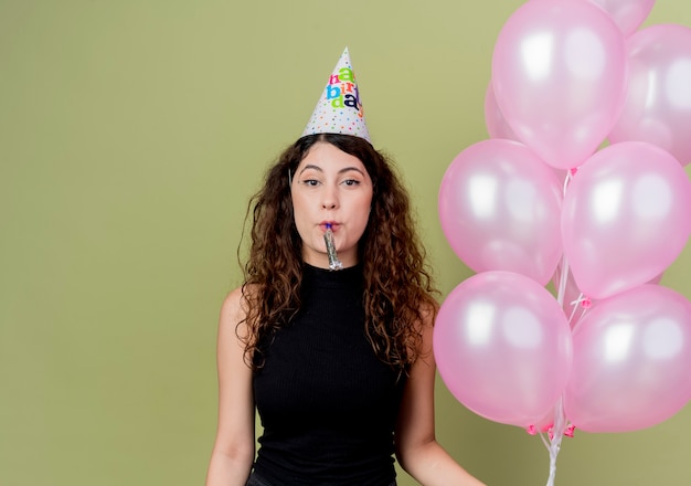 Молодая красивая женщина с вьющимися волосами в праздничной шапочке держит воздушные шары, дует в свисток, счастливая и позитивная, празднует день рождения, стоя над светлой стеной