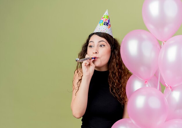 Young beautiful woman with curly hair in a holiday cap holding air balloons blowing whistle happy and positive celebrating birthday party standing over light wall