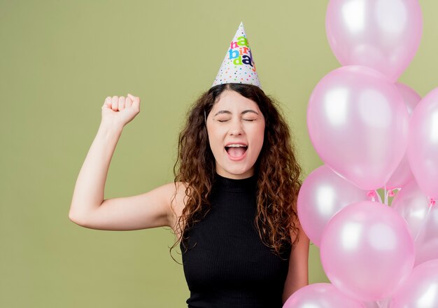 Young beautiful woman with curly hair in a holiday cap holding air balloons clenching fist crazy happy birthday party concept standing over light wall