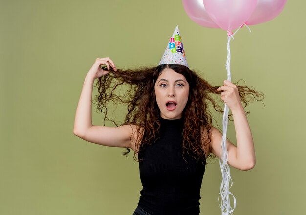 Free photo young beautiful woman with curly hair in a holiday cap holding air balloons  confused and surprised birthday party concept standing over light wall