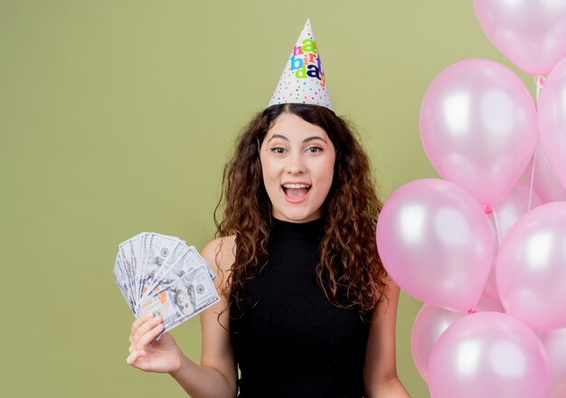 明るい壁の上に立っている現金の誕生日パーティーのコンセプトを示して幸せで興奮している気球を保持しているホリデーキャップで巻き毛の若い美しい女性