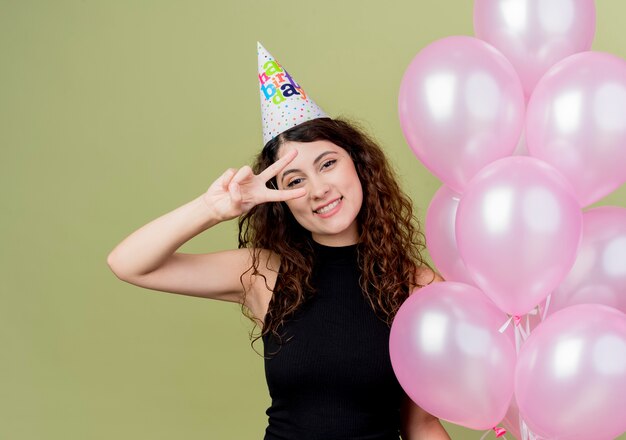 明るい壁の上に立っている誕生日パーティーを祝うvサインを示す幸せでポジティブな気球を保持しているホリデーキャップで巻き毛の若い美しい女性