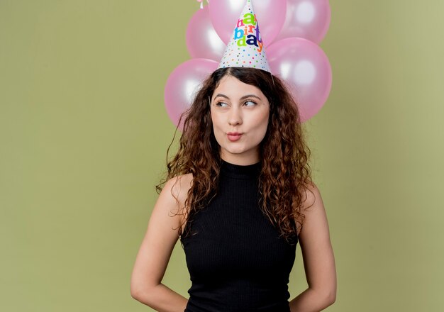 明るい壁の上に立って幸せで前向きな誕生日パーティーのコンセプトを脇に見て気球を保持しているホリデーキャップで巻き毛の若い美しい女性