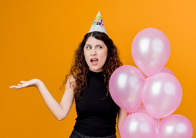 Young beautiful woman with curly hair in a holiday cap holding air balloons looking confused birthday party concept standing over orange wall