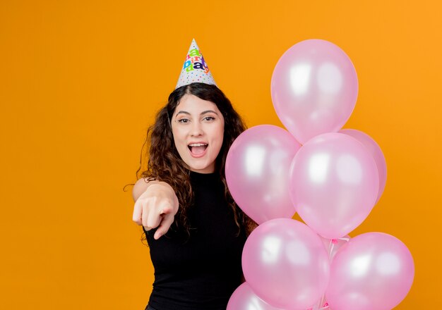 Молодая красивая женщина с вьющимися волосами в праздничной кепке держит воздушные шары, указывая пальцем на камеру, весело улыбаясь концепции вечеринки по случаю дня рождения над оранжевым