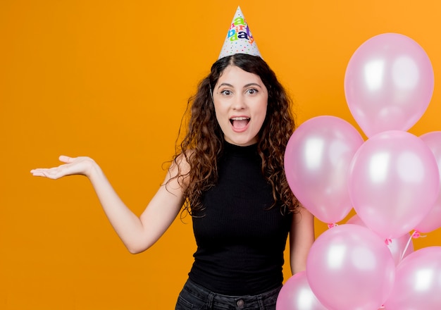 Giovane bella donna con capelli ricci in un cappello di vacanza che tiene gli aerostati di aria che presentano qualcosa con il braccio della mano concetto di festa di compleanno felice ed emozionato sopra l'arancio