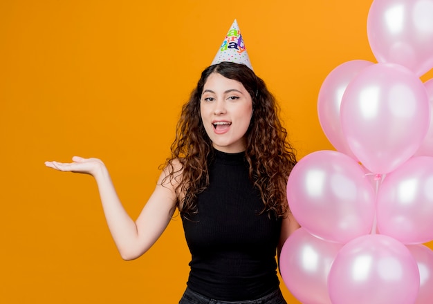 Giovane bella donna con i capelli ricci in un cappello di vacanza che tiene gli aerostati di aria che presentano qualcosa con il braccio che sorride allegramente concetto di festa di compleanno in piedi sopra la parete arancione