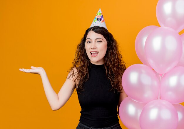 Young beautiful woman with curly hair in a holiday cap holding air balloons presenting something with arm smiling cheerfully birthday party concept standing over orange wall