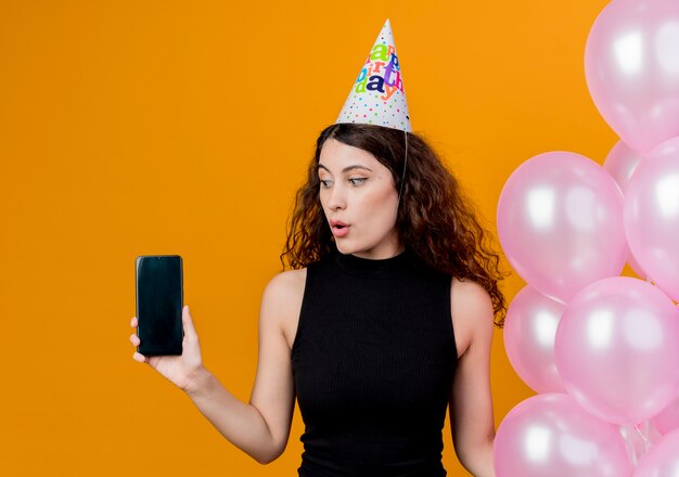 オレンジ色の壁の上に立っている混乱した誕生日パーティーのコンセプトを見てスマートフォンを示す気球を保持しているホリデーキャップで巻き毛の若い美しい女性