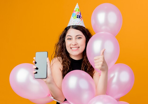 オレンジ色の上に幸せで興奮した誕生日パーティーのコンセプトを親指を示す気球とスマートフォンを保持しているホリデーキャップで巻き毛の若い美しい女性