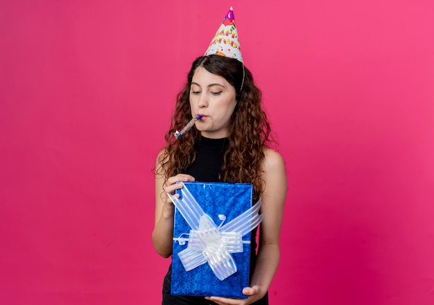 Foto gratuita giovane bella donna con capelli ricci in una protezione di vacanza che tiene la scatola di compleanno che soffia il concetto di festa di compleanno del fischio che sta sopra il muro rosa