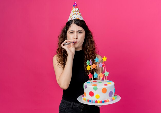 ピンクの壁の上に立って幸せでポジティブな誕生日パーティーのコンセプトを吹く誕生日ケーキを保持しているホリデーキャップで巻き毛の若い美しい女性