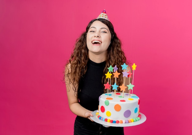 Young beautiful woman with curly hair in a holiday cap holding birthday cake happy and excited smiling cheerfully birthday party concept  over pink