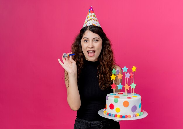 ピンクの壁の上に立っている誕生日ケーキ幸せで興奮した笑顔の誕生日パーティーのコンセプトを保持しているホリデーキャップで巻き毛の若い美しい女性