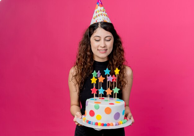 핑크를 통해 생일 케이크 행복하고 긍정적 인 생일 파티 개념을 들고 휴가 모자에 곱슬 머리를 가진 젊은 아름 다운 여자