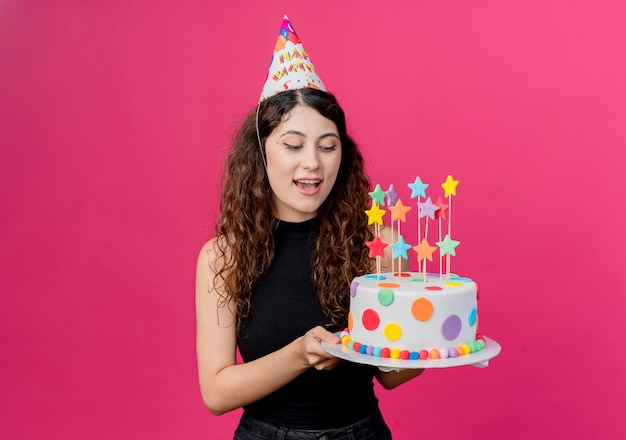 Young beautiful woman with curly hair in a holiday cap holding birthday cake happy and positive birthday party concept  over pink