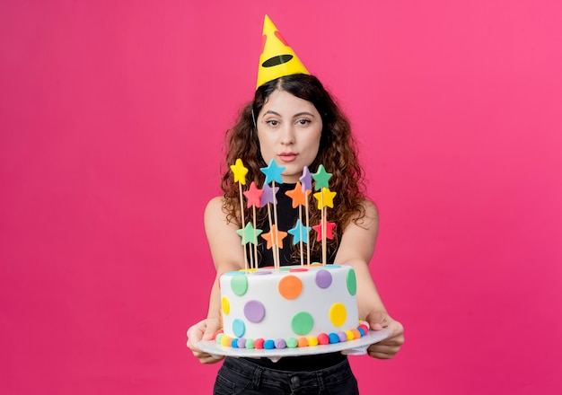 분홍색 벽 위에 생일 케이크 행복하고 긍정적 인 생일 파티 개념 서 들고 휴가 모자에 곱슬 머리를 가진 젊은 아름 다운 여자